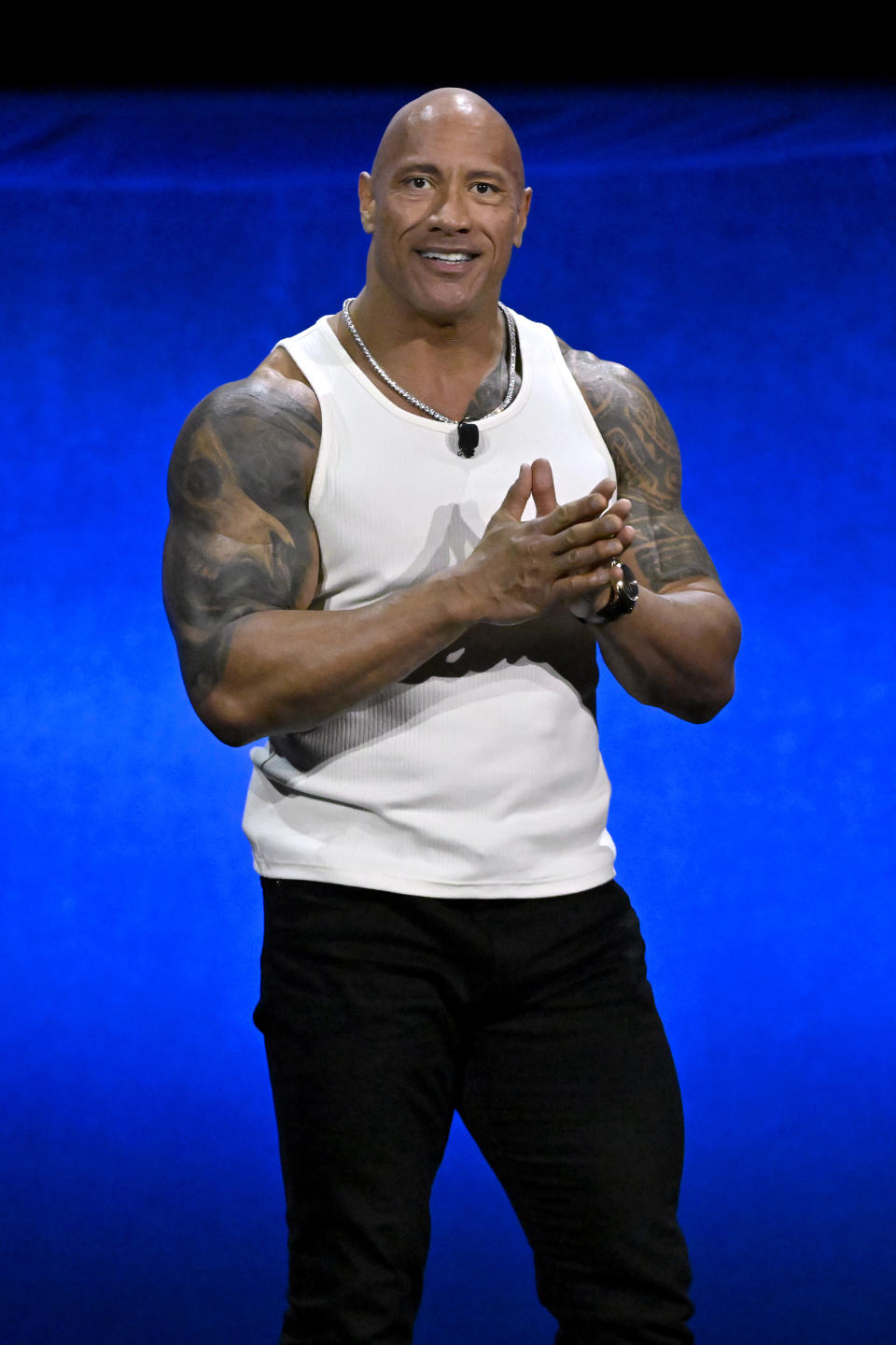 Dwayne "The Rock" Johnson stands with clasped hands, wearing a sleeveless white shirt and black pants, showcasing his tattooed arms