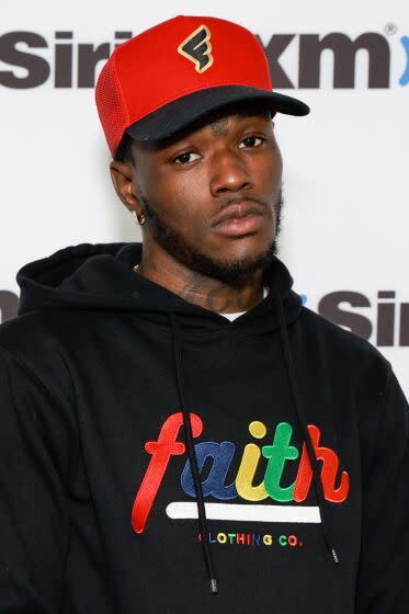 DC Young Fly is posing with a serious face while wearing a red baseball cap and a black hoodie with multi-color "faith"