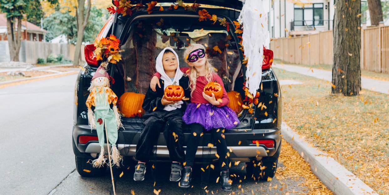 boy and girl holding jack o lanterns in open tailgate of parked vehicle with leaves blowing and on bunting at top of tailgate there is a fabric scarecrow on a stick leaning against vehicle the girl is wearing a black mask and purple tutu