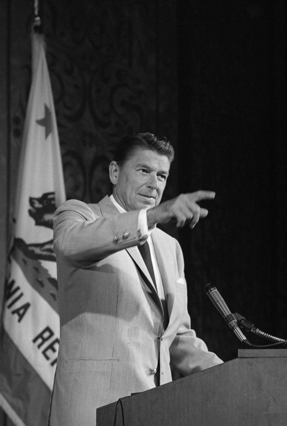 ronald reagan pointing as he stands at a podium with a california flag behind him