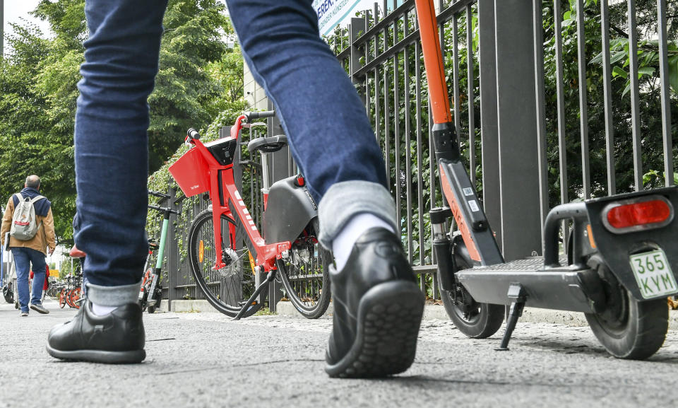 Numerous e-bikes and e-scooters stand on a footpath where pedestrians are on the move. | Jens Kalaene—Jens Kalaene/picture-alliance/dp/AP