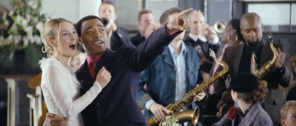 Juliet and Peter, in a white dress and a suit, stand in the aisle of a church pointing and exclaiming - two men with saxophones are seen behind them