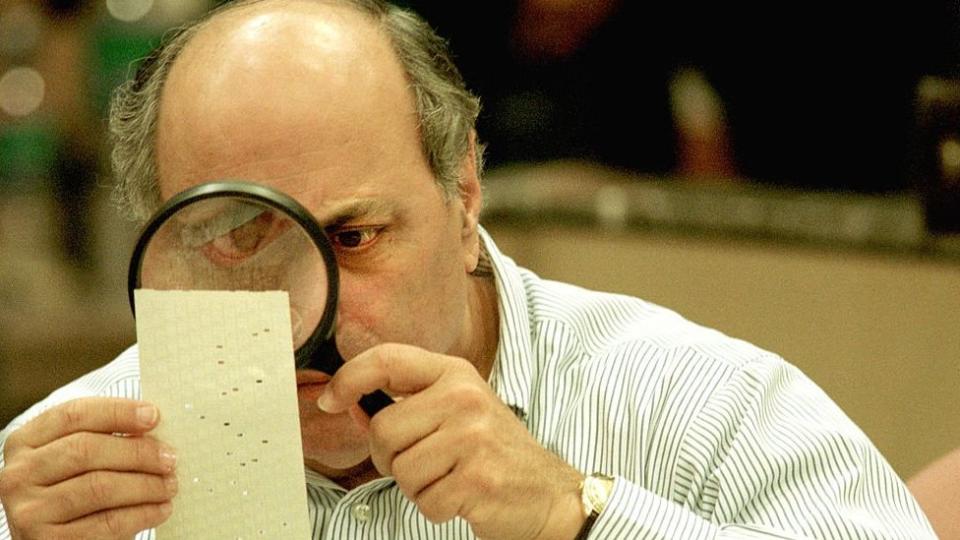 Judge Robert Rosenberg of the Broward County Canvassing Board uses a magnifying glass to examine a dimpled chad on a punch card ballot November 24, 2000 during a vote recount in Fort Lauderdale, Florida. On May 4, 2001