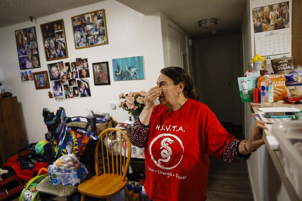 A woman in a red T-shirt holds a hand to her eyes inside a home with pictures on the wall and furnishings