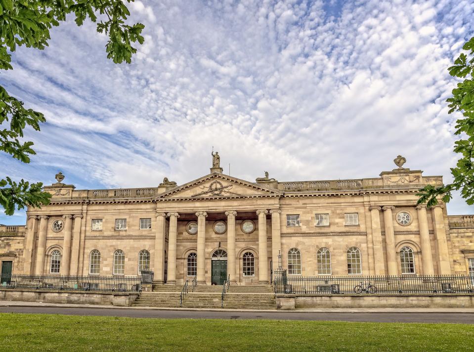 18th Century architecture of York County Court.  There is a carving on the cornice and a statue above.  A dramatic sky is above and the view is framed with foliage.