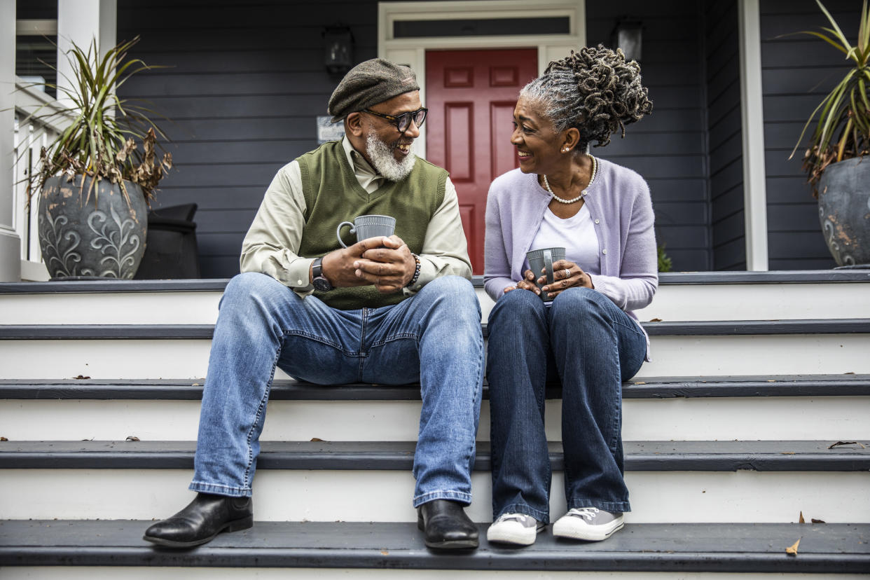 An older man and an older woman hold coffee mugs as they sit on the front steps of a house.