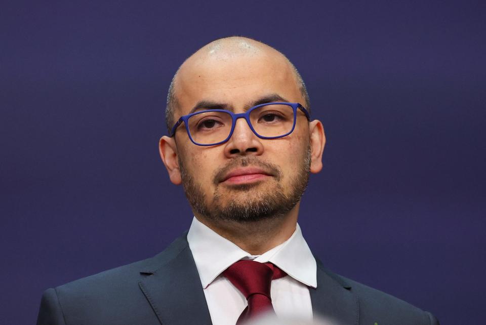 Demis Hassabis wearing a suit, a red tie, and blue-framed glasses