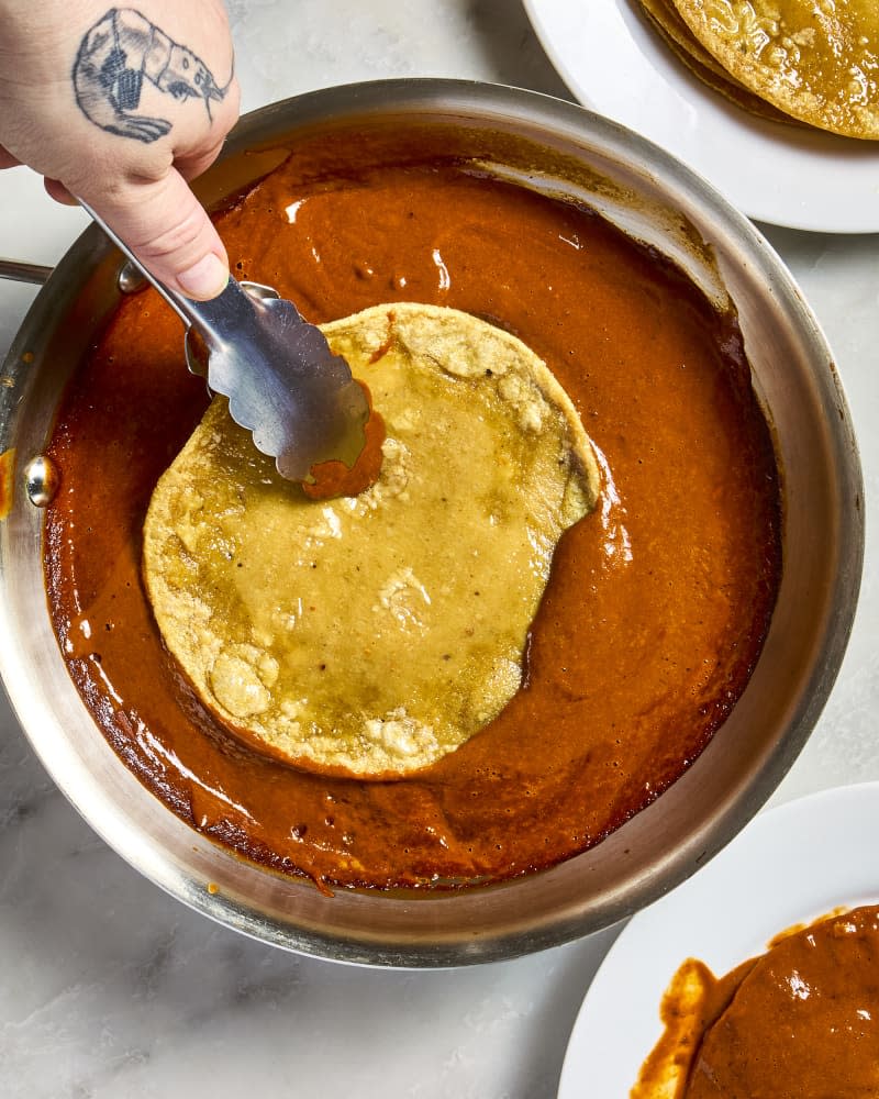 A hand dipping a tortilla into enchilada sauce with tongs