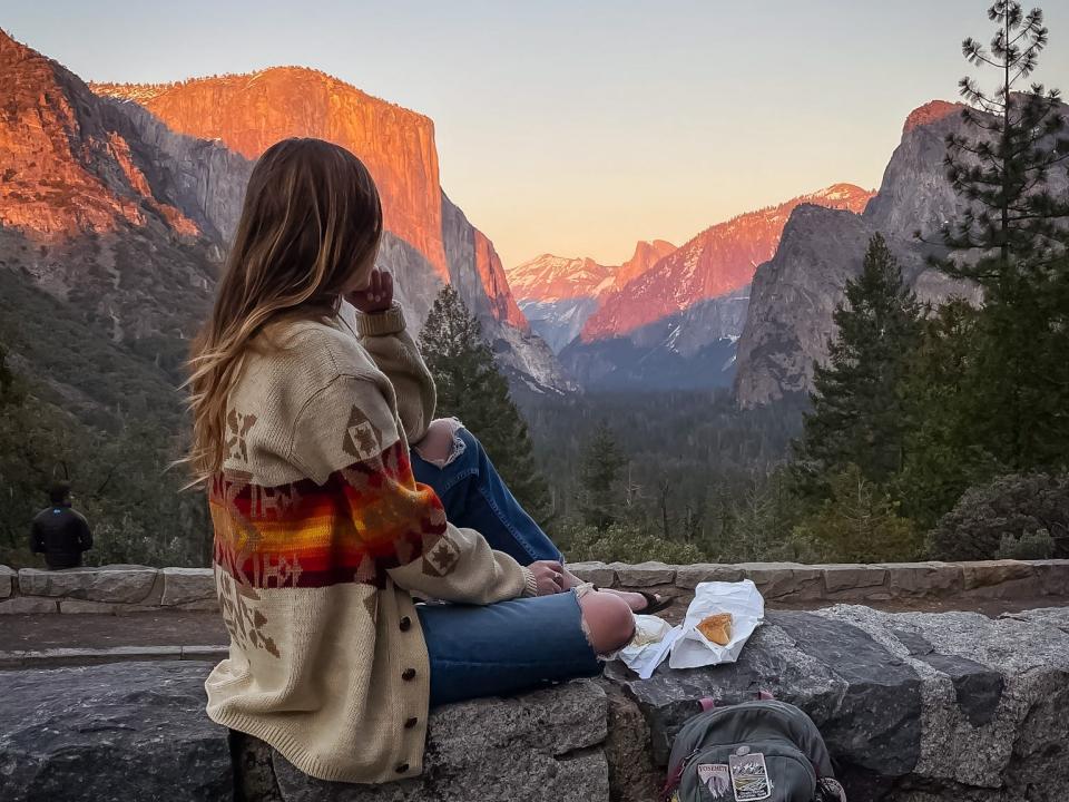 Emily, with food and a backpack covered in patches next to her, sitting on a wall looking out at the mountains at Yosemite National Park.