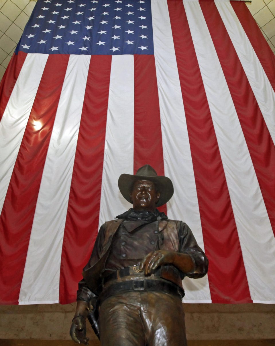 FILE - In this Sept. 11, 2011, file photo, a bronze statue of late actor John Wayne stands before a four-story high United States flag at John Wayne Orange County Airport in Santa Ana, Calif. In the latest move to change place names in light of U.S. racial history, leaders of Orange County’s Democratic Party are pushing to drop film legend Wayne’s name, statue and other likenesses from the county’s airport because of his racist and bigoted comments. (AP Photo/Reed Saxon, File)