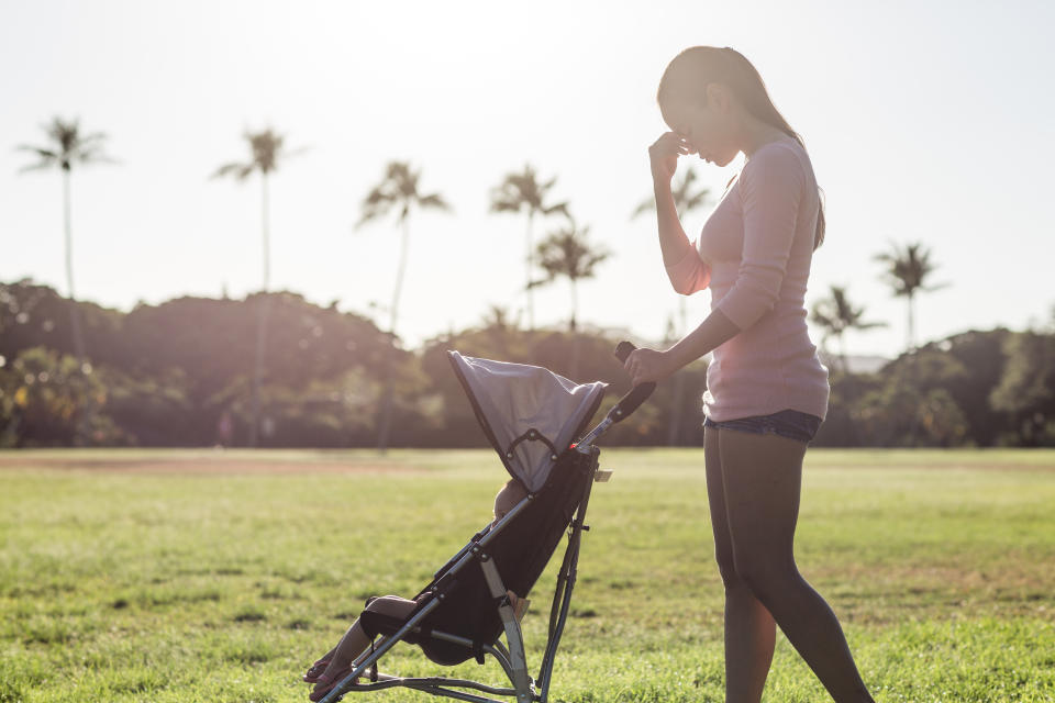 Sleepless single female parent with hand on her face. She is pushing a stroller with her baby child at the park during a sunny summer day. Maternity stress.