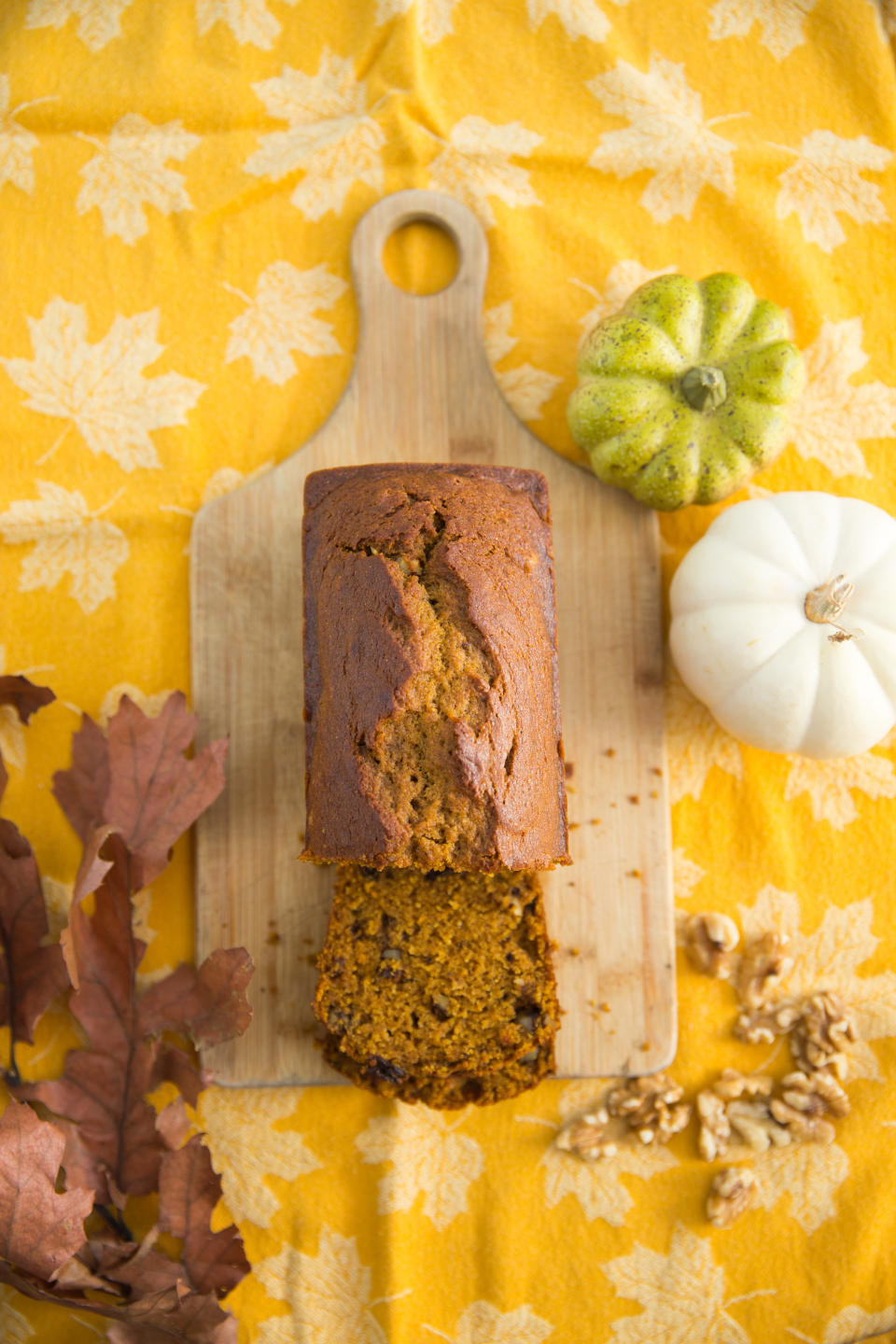 A loaf of pumpkin bread with a slice cut, displayed on a wooden board surrounded by nuts, leaves, and decorative gourds on a patterned cloth