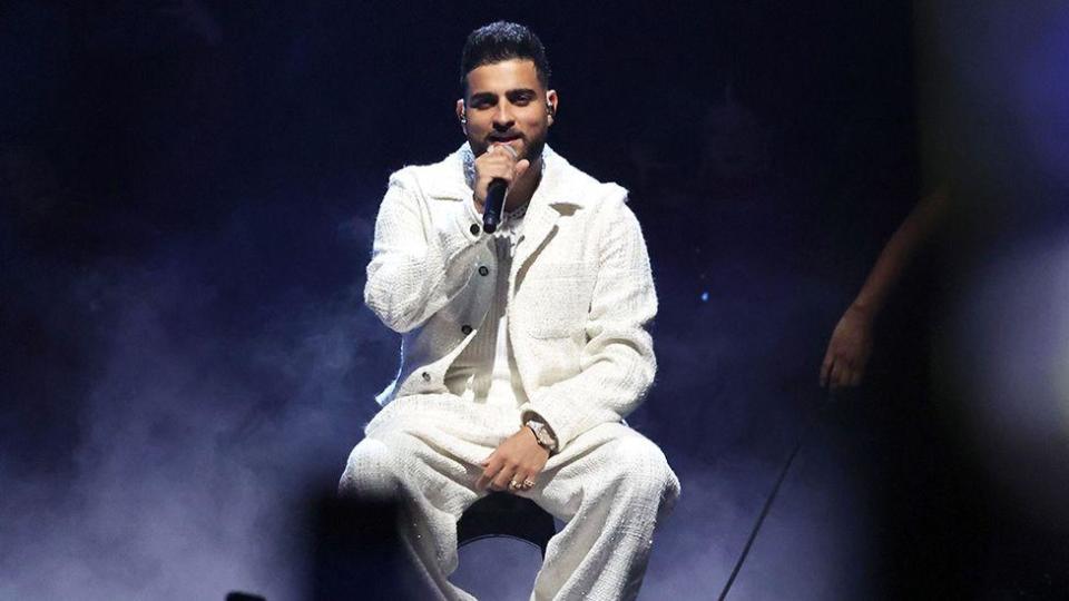 A man wearing all white sitting on stage with a mic in his hand, wearing all white. The background is dark and smoky.