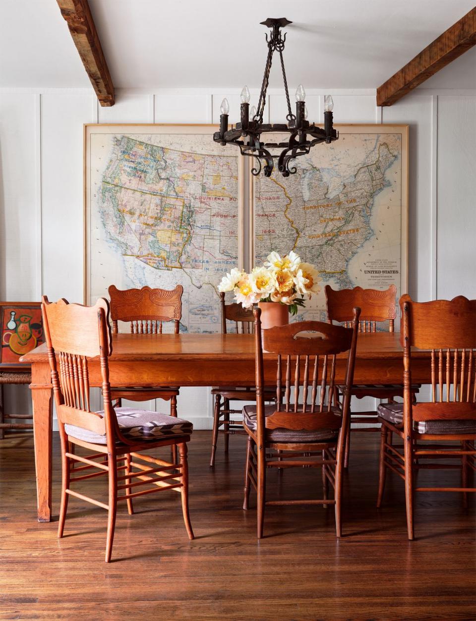 long wood dining table with a vase of flowers, six press back oak chairs with fabric seat cushions, framed wall size antique map of the united states, cast iron pendant with teardrop bulbs, wood floor, beamed ceiling