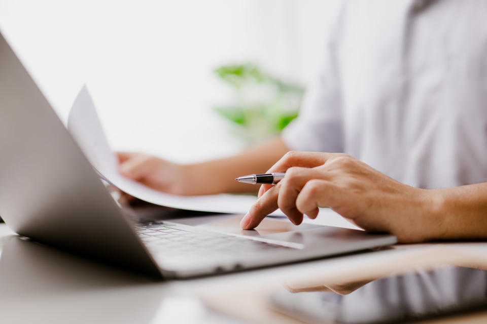 A person types on their laptop while holding a document and pen in their hands