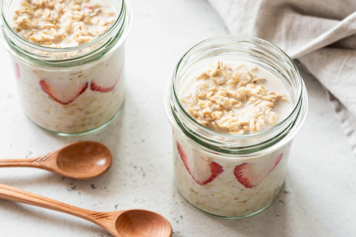 Two jars of overnight oats with strawberries in glass jars with two wooden spoons, on a speckled white table with a beige napkin