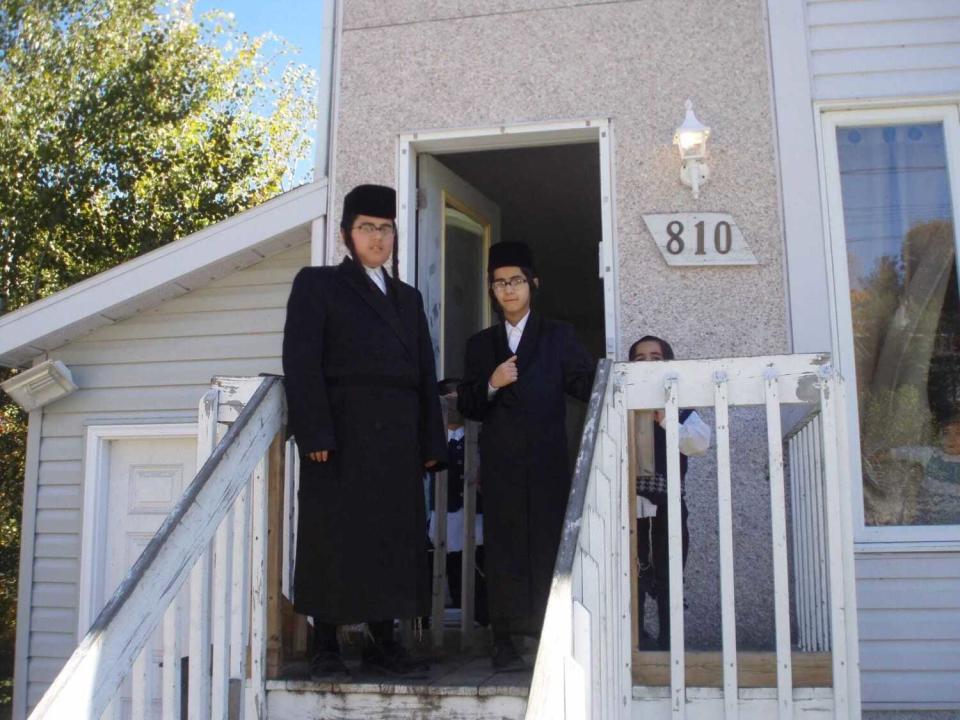 Two people with dark clothing, hats and earlocks stand near stairs outside a home