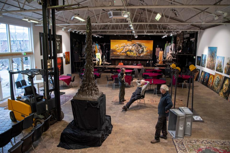 One man sits while two others stand in an art gallery space featuring the Pillar of Shame sculpture.