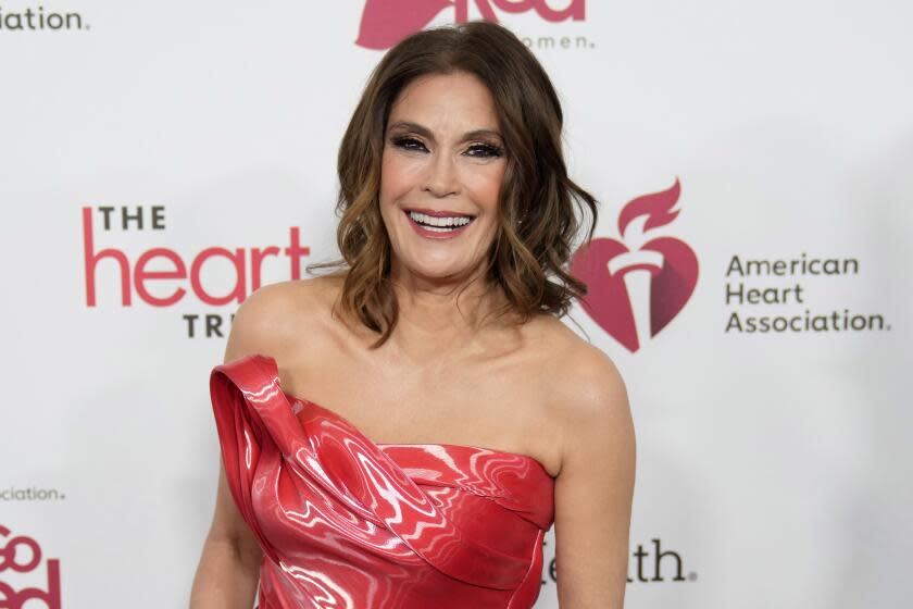 A woman with wavy shoulder-length brown hair in a shiny, strapless red dress smiling against a white background