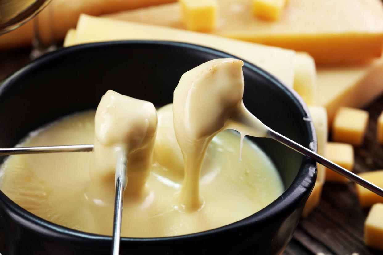 Gourmet Swiss fondue dinner on a winter evening with assorted cheeses on a board alongside a heated pot of cheese fondue with two forks dipping bread and white wine behind in a tavern or restaurant