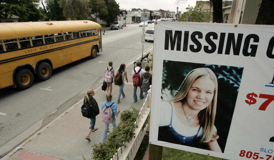 A "Missing" billboard above a sidewalk as young people and a school bus go by