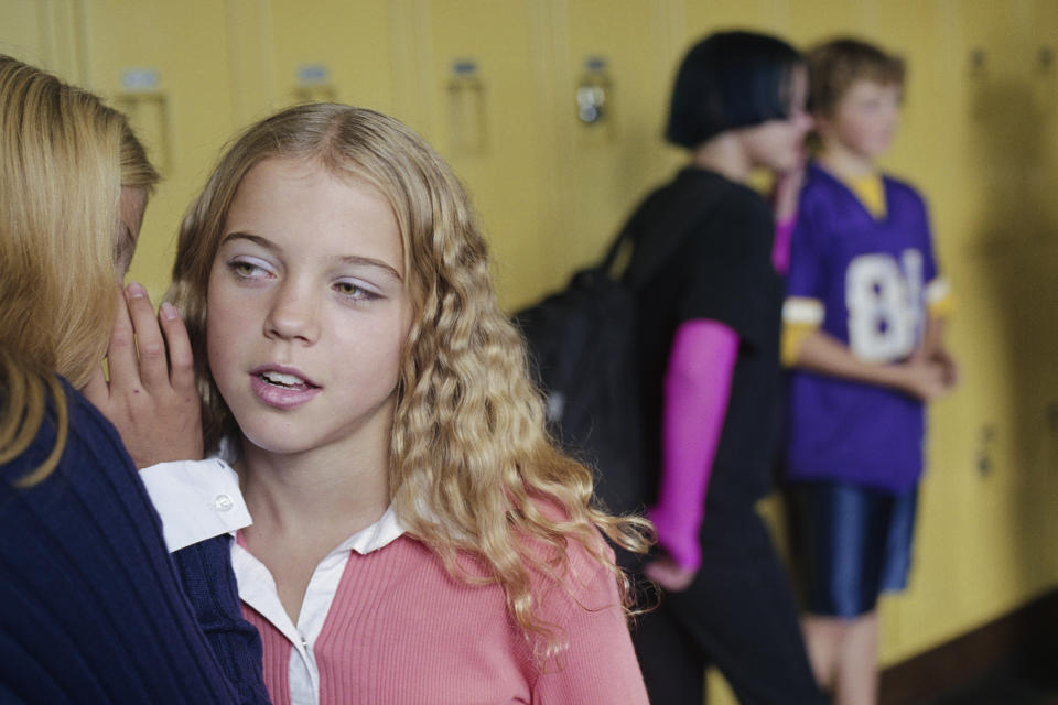 Two girls whisper in a school hallway while two other students stand by lockers in the background