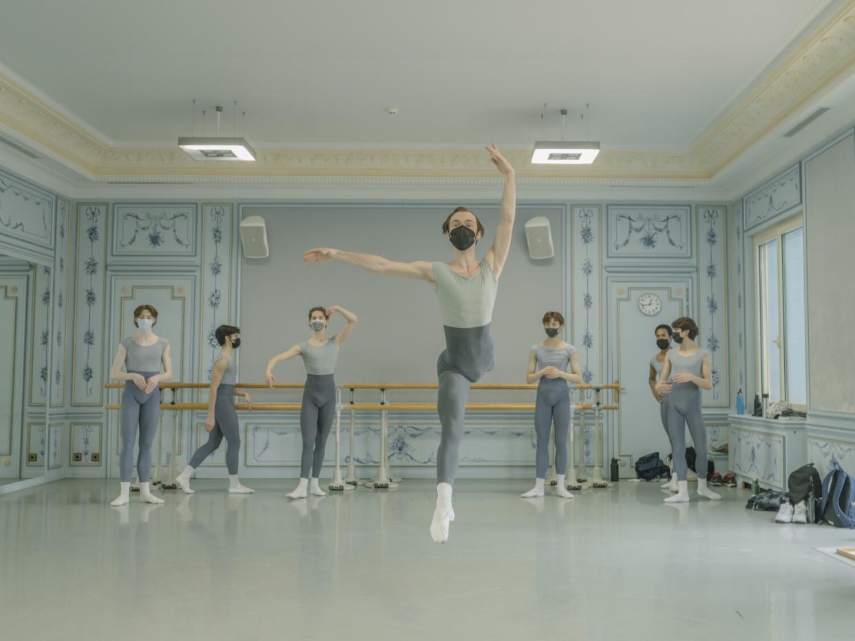 A boy leaps in a ballet studio with other boys at the bar behind him