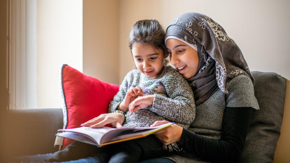 A pre-teen girl wearing a hijab sits on a couch with her little sister on her lap and reads her a bedtime story.