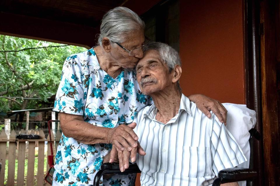 old couple, woman standing kissing head of man sitting down, eyes closed 