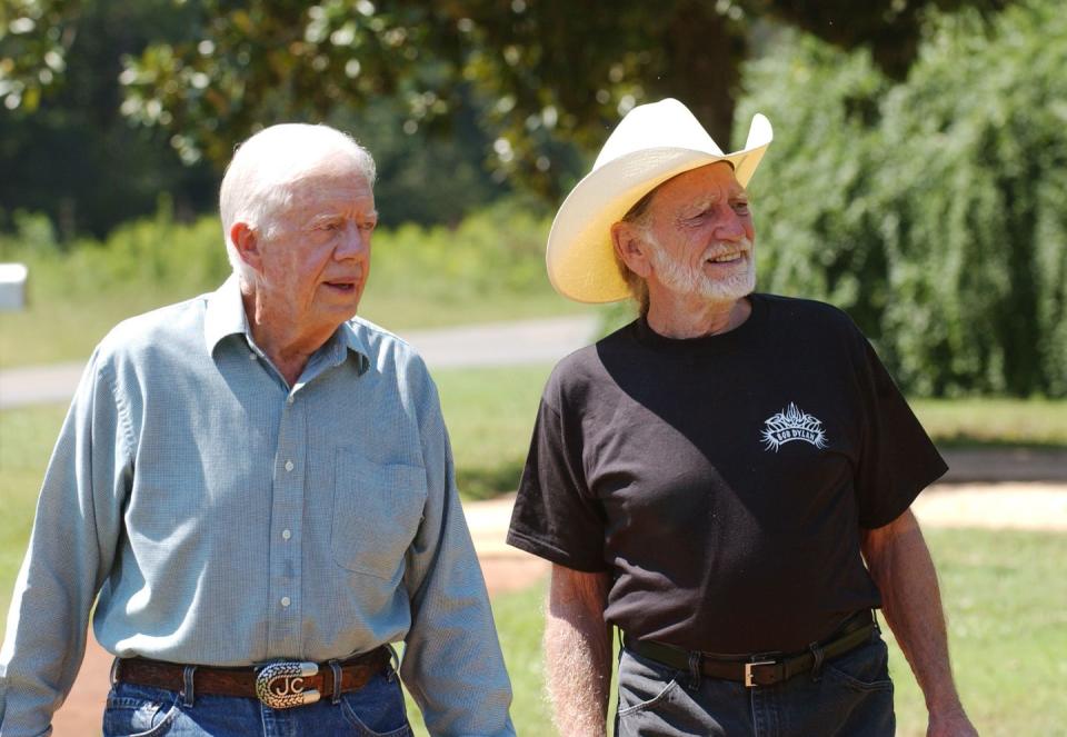 jimmy carter, wearing a blue shirt, and willie nelson, wearing a black t shirt and cowboy hat, walking together outside