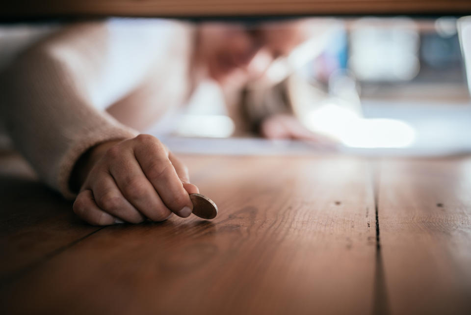 Person reaches under furniture to pick up a small coin from the wooden floor; face blurred and not fully visible in the background