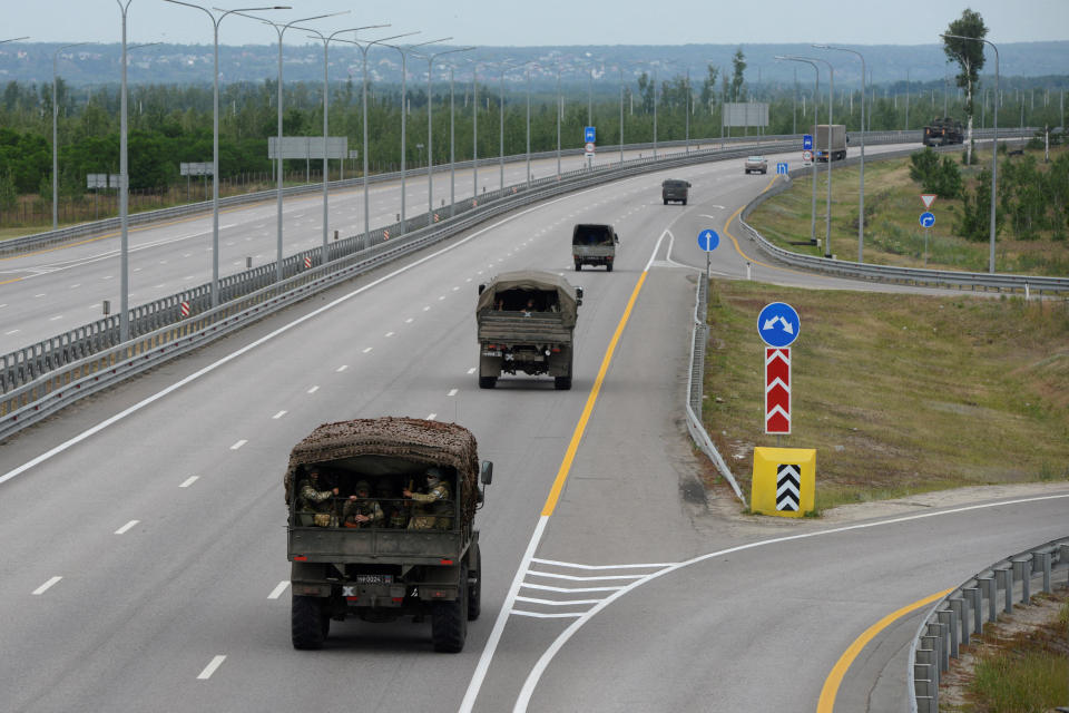 A half dozen military vehicles with soldiers aboard drive in a convoy spaced apart on what appears to be a closed highway.