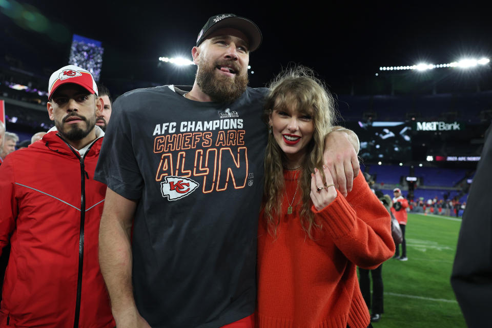 Travis Kelce in a Chiefs t-shirt hugs Taylor Swift wearing a red sweater