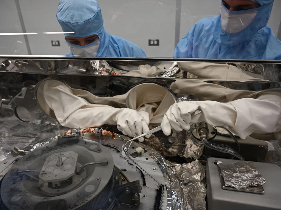 Two people in blue protective gear and white gloves use tools on the container holding the Bennu asteroid sample