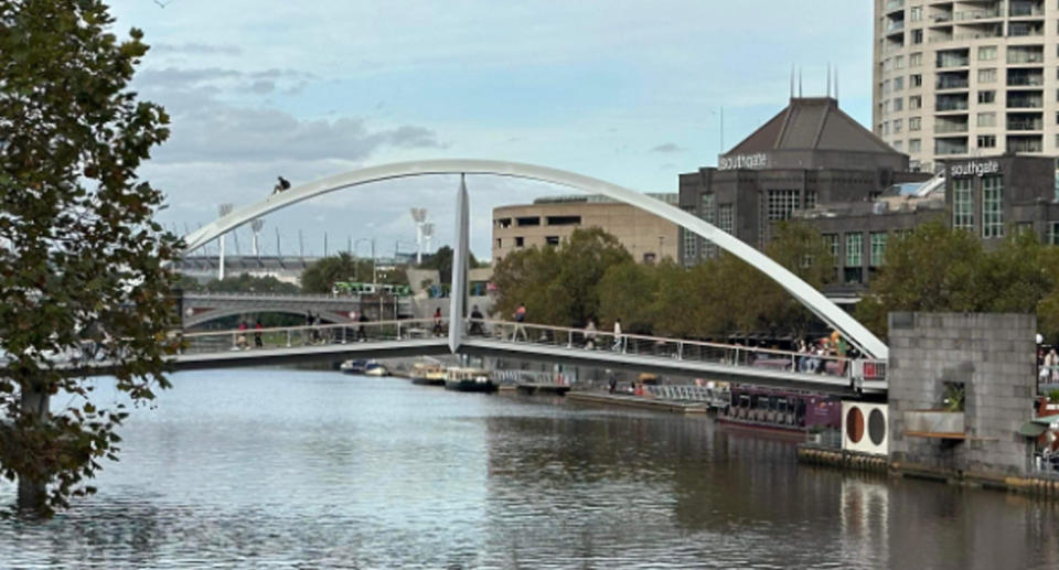 The man can be seen sitting on the bridge's arch as he slide down the bridge, with commuters walking below. 