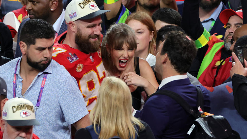 Taylor Swift and Travis Kelce smile in a crowd of people.