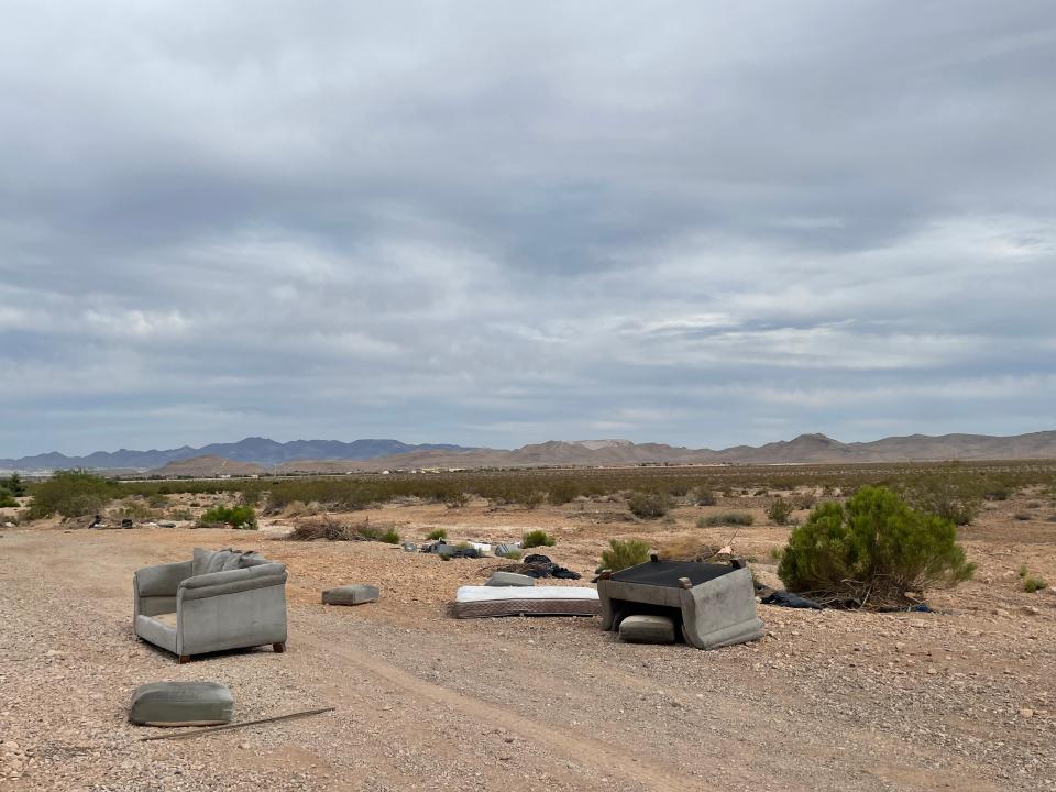 Two couches, a mattress, and other trash laying on the side of a dirt road in the desert.