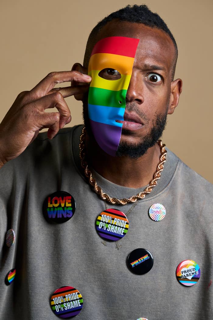 Marlon Wayans holding a rainbow mask halfway over his face, wearing a thick gold chain and a sweater adorned with pro-LGBTQ+ buttons