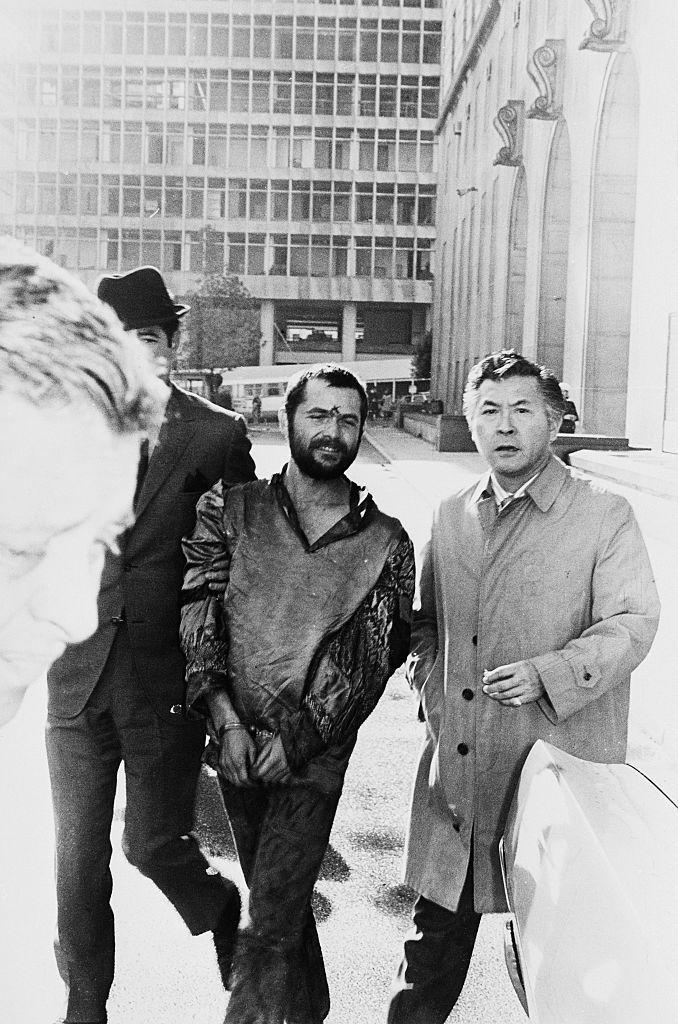 a man with an x carved in his forehead smiles as he is led away from a courthouse in handcuffs next to a man in a trenchcoat