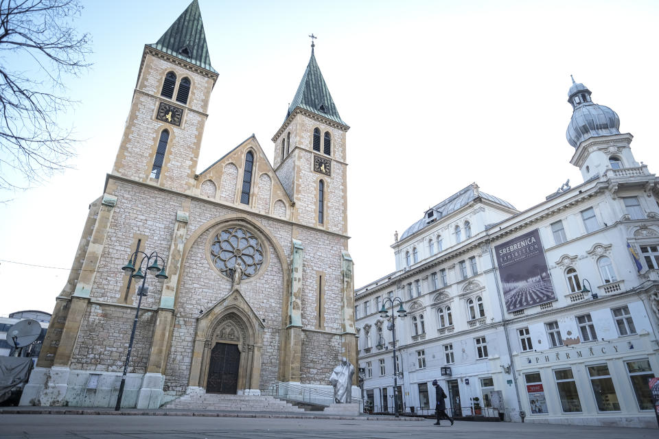 In this Friday, April 10, 2020 photo, a person wearing a face mask walks on the deserted streets around the Sacred Heart Cathedral in Sarajevo, Bosnia, during the national lockdown the authorities have imposed attempting to limit the spread of the new coronavirus. Traditionally, hundreds, sometimes thousands of residents who fled during the 1990s war and the years of economic instability that followed, return to Sarajevo each spring to celebrate religious or cultural holidays with family and friends. This year, nobody is returning leaving the streets and places for religious gatherings empty as families and friends celebrate the holidays apart due to the COVID-19 lockdown. (AP Photo/Kemal Softic)