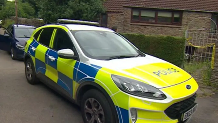 A picture of a police car at the scene of the incident. A brown brick house is visible in the background, with yellow police tape seen hanging from the gate. 