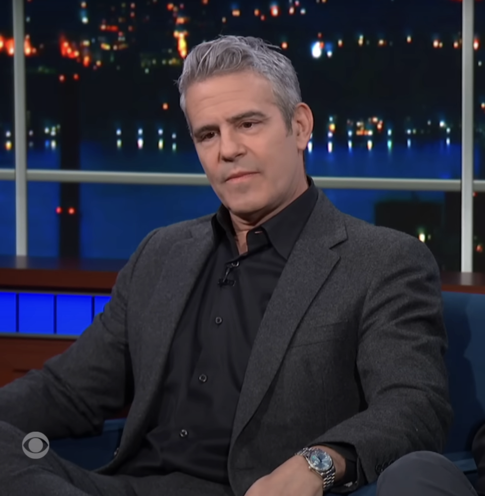 Andy in a talk show setting, wearing a dark suit and sitting with crossed legs