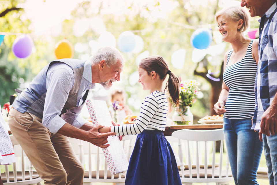 Elderly man presents a gift to a young girl as middle-aged woman and man look on, with party balloons hanging in the background