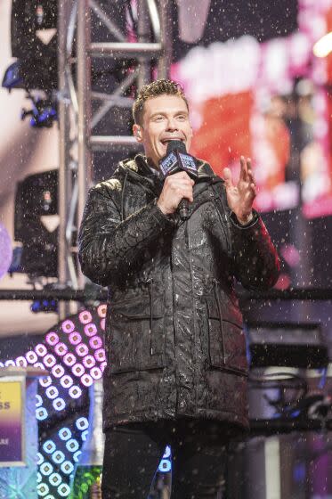 Ryan Seacrest in a black puffer jacket speaking into a microphone in the snow