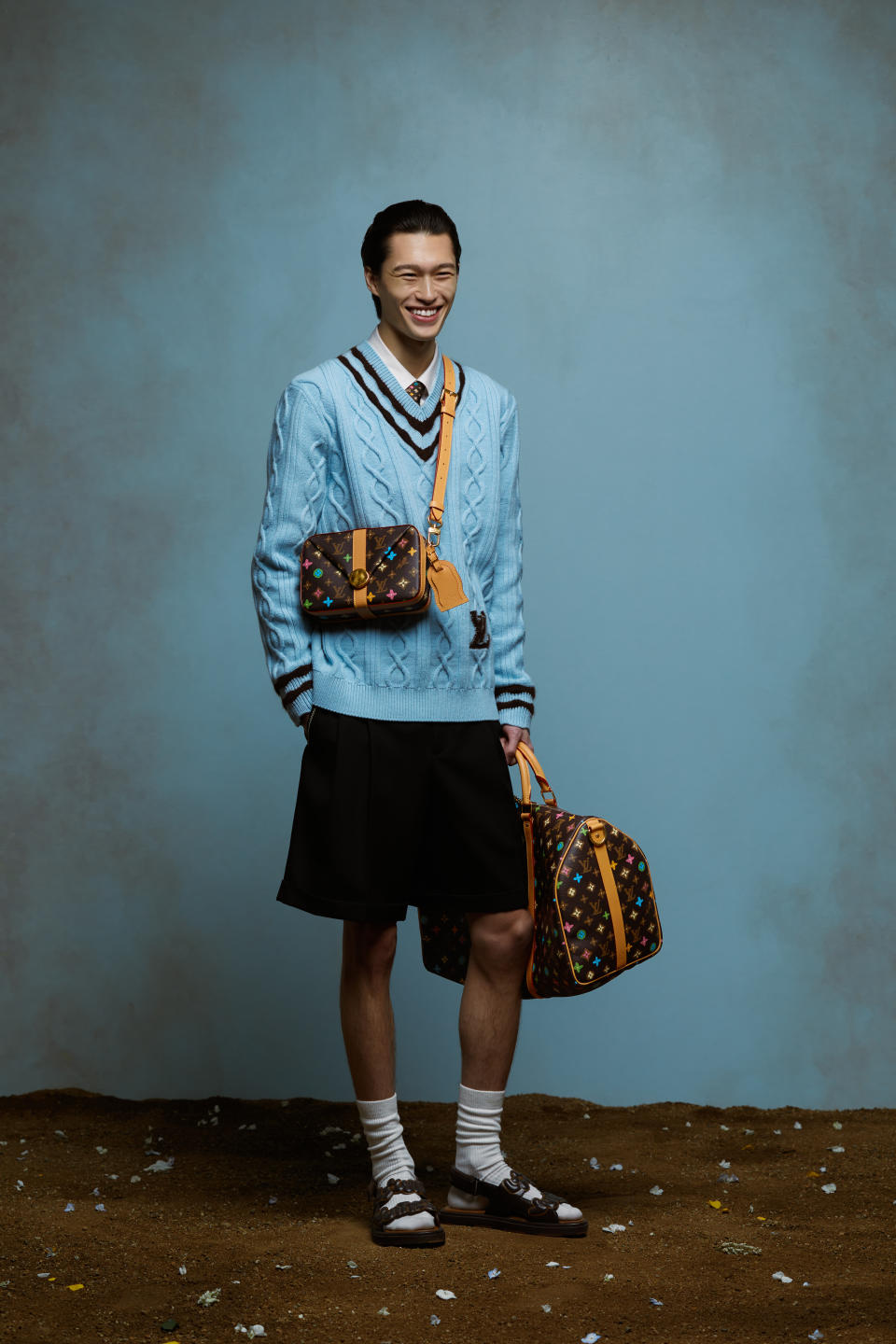 A model wears a baby blue cable knit jumper and dark shorts, together with bags printed with a Craggy Monogram pattern, from the Louis Vuitton spring 2024 men's capsule collection by Tyler, the Creator. 