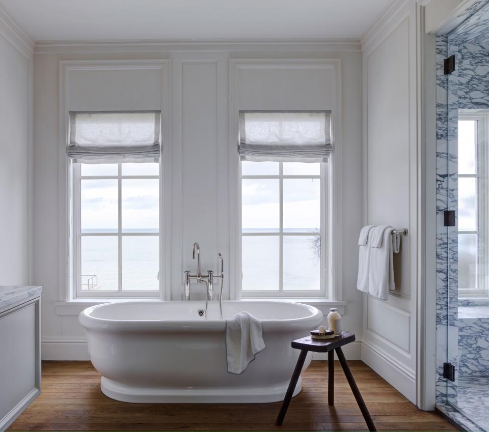 Honed Arabescato Corchia marble in the walk-in shower breaks up the monochromatic white of the master bath’s paneled walls. A tub by Waterworks overlooks Lake Michigan through windows dressed in Roman shades made with De La Cuona cashmere.