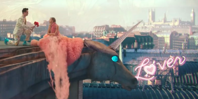 A screenshot from the "ME!" video, Taylor sits on the edge of a building with a city in the background, a bright pink neon sign reads Lover on a rooftop