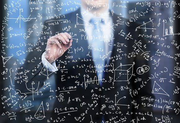 A man in a suit writes formulas on a glass wall in front of him.