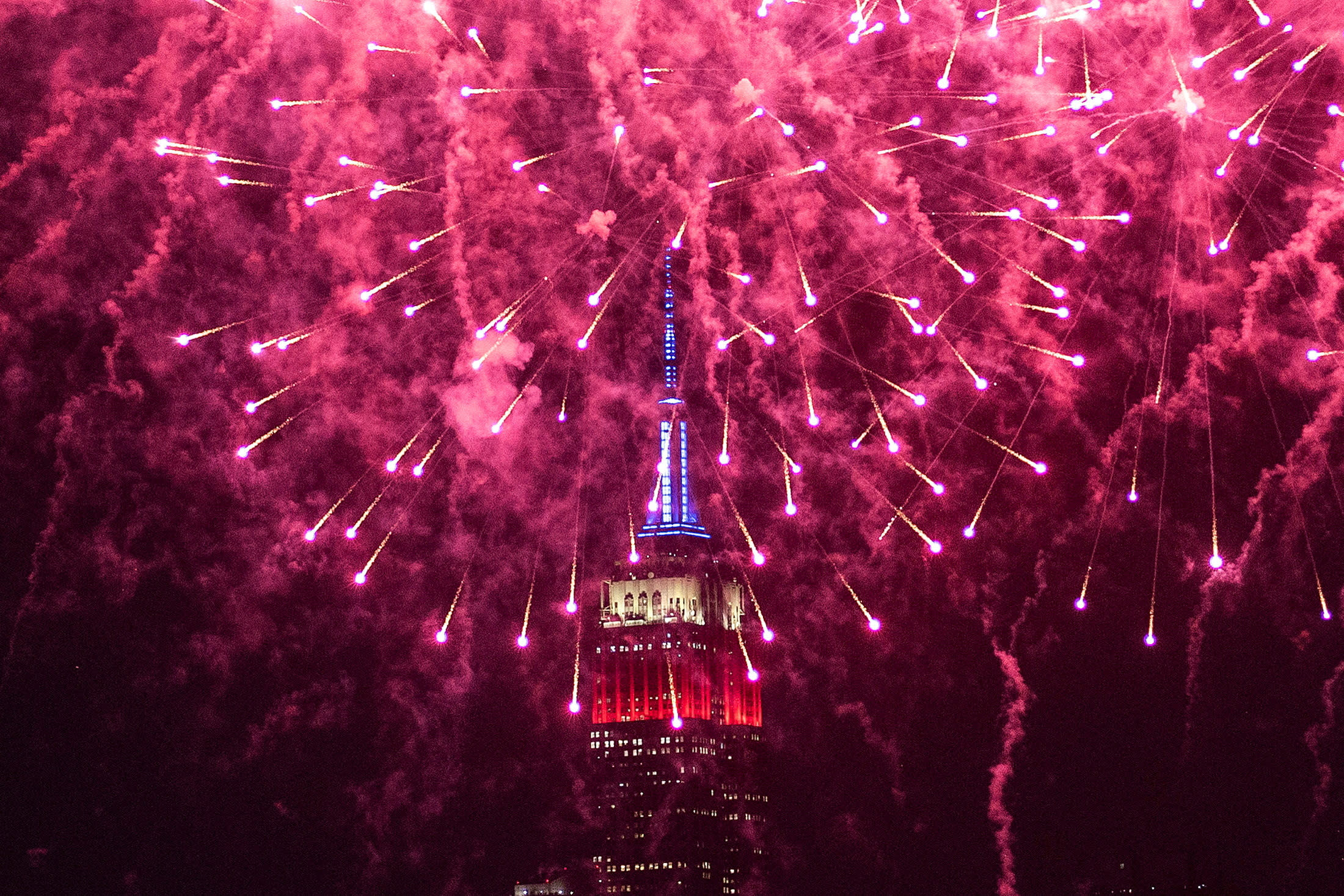 A view of red fireworks lighting up the sky over the Empire State Building, which is lit up in red, white and blue.