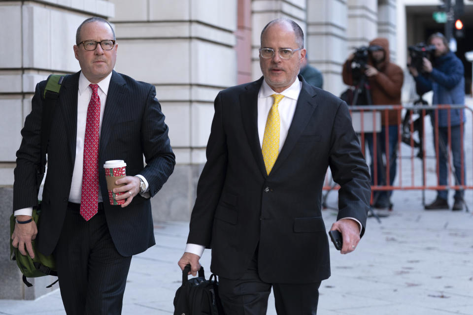 James Lee Bright and Phillip Linder, Bright holding a coffee cup and a bag over his shoulder, and Linder carrying a briefcase, walk past a barricade and into the courthouse in suits and ties.